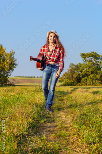 Beautiful, happy, red-haired girl in a field on a sunset background with a guitar in the summer. Concept of inspiration by nature