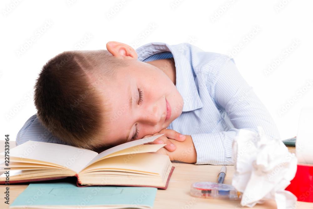 Cute little school boy with huge smile sitting at his desk on white background. Happy intelligent children in shirt with blue eyes, lies on table with book and sleep Cute little school boy with huge smile sitting at his desk on white background. Happy intelligent children in shirt with blue eyes, lies on table with book and sleep