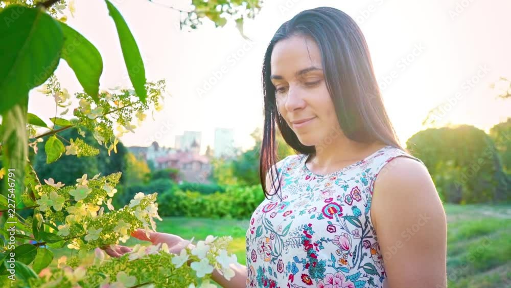 Portrait of a smiling brunette with dimpled cheeks against a green park. Beautiful young woman stands in park and looks at camera. Pretty girl in summer dress with floral print Portrait of a smiling brunette with dimpled cheeks against a green park. Beautiful young woman stands in park and looks at camera. Pretty girl in summer dress with floral print