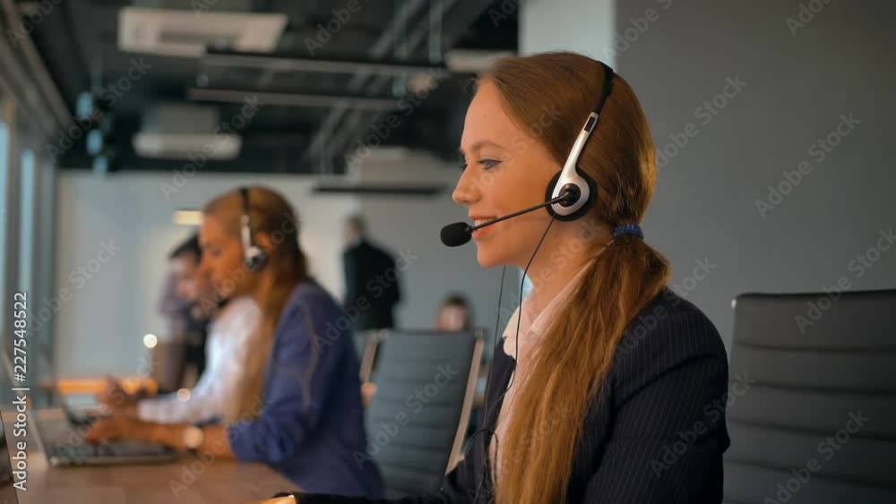 Call-center employees work in deal office. Young handsome female in headset on first plan talking with customer. Modern business room with beautiful view. 4k UltraHD.