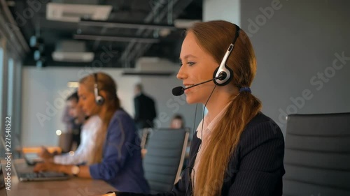 Call-center employees work in deal office. Young handsome female in headset on first plan talking with customer. Modern business room with beautiful view. 4k UltraHD.