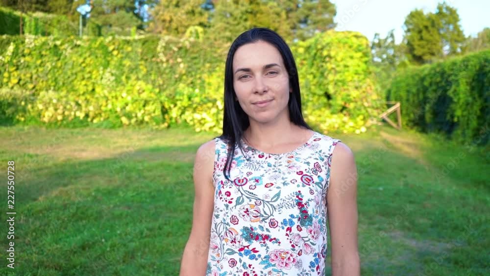 Portrait of a smiling brunette with dimpled cheeks against a green park. Beautiful young woman stands in park and looks at camera. Pretty girl in summer dress with floral print Portrait of a smiling brunette with dimpled cheeks against a green park. Beautiful young woman stands in park and looks at camera. Pretty girl in summer dress with floral print