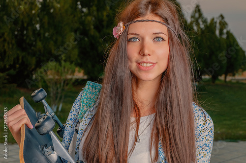 Lovely girl in boho attire with skatebord and artificial flower in her hairs looking at camera and smiling photo