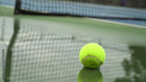 Abandoned tennis ball sitting in a shallow puddle courtside.  Blue and green surfaced court.  Cloudy, cold day.  Net reflected in the puddle surface.