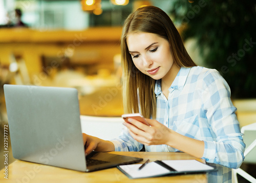 Young beautiful blond girl sitting in a coffee shop and working on computer. Image of happy woman using laptop and smartphone in cafe.