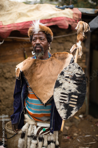 Traditional tribal African man dressed in animal skins and carrying a shield standing outside his homestead photo