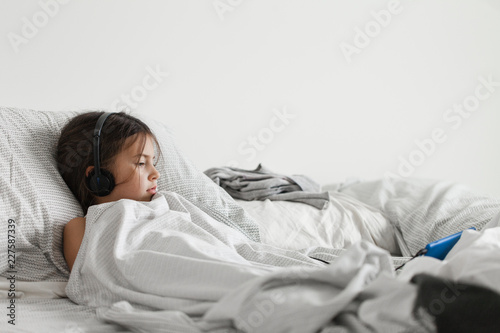 Girl laying in bed with the covers pulled up while watching an electronic device with headphones
