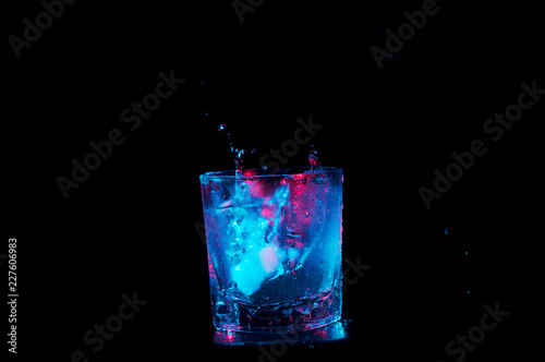 Ice cubes splashing into a rocks glass of liquid under colorful lights isolated on a black background