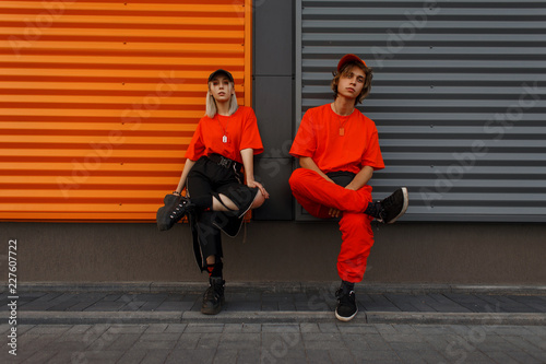 Beautiful fashionable young stylish couple in fashion orange clothes with caps sit near the metal gray and orange wall
