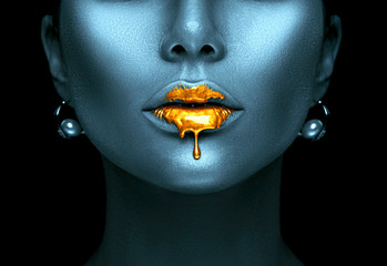Gold paint drips from the sexy lips, golden liquid drops on beautiful model girl's mouth, creative abstract dark blue skin makeup. Beauty woman face