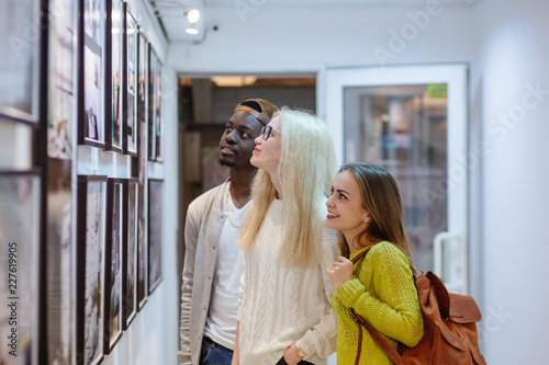 Fényképezés Three young people hipster students with different skins looking happy together, talkng, looking at picture on wall, spending free time together in art photo gallery