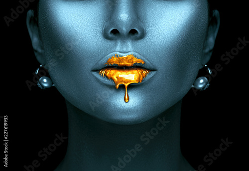 Gold paint drips from the sexy lips, golden liquid drops on beautiful model girl's mouth, creative abstract dark blue skin makeup. Beauty woman face