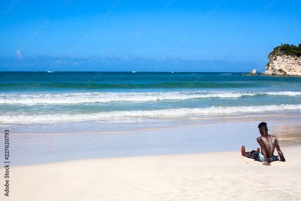 Black man sits on the beach on white sand, on a blue sea with waves and sky background