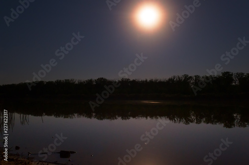 the stars in the night sky are reflected in the mirror smooth surface of the lake, and the moon illuminates the forest on the opposite bank