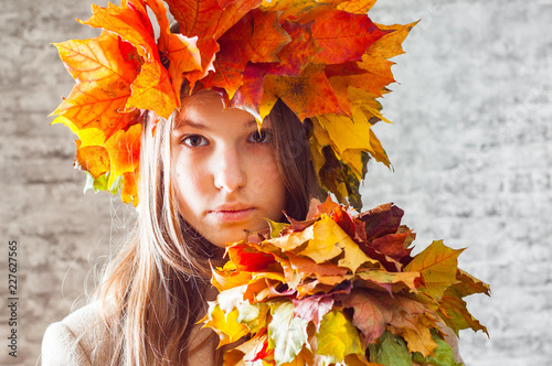portrait of young teenager brunette girl with long hair with a wreath of autumn leaves on her head on gray wall background