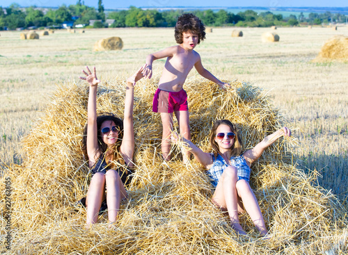 Two elder sisters and a little brother sitting on a haystack in the field on a sunny summer day. Showing affection and warm relationships concept