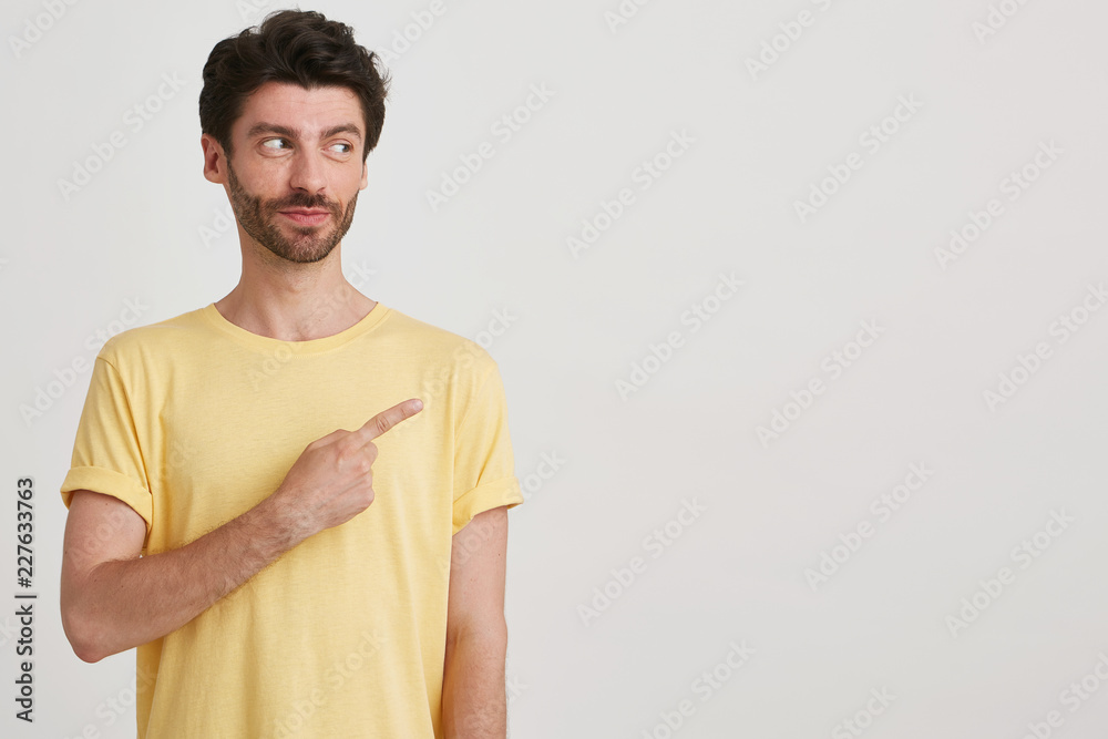 Close up of attractive young man with restrained smile, dark brown hair and beard wears yellow casual tshirt, points to the right side with finger isolated over white background Close up of attractive young man with restrained smile, dark brown hair and beard wears yellow casual tshirt, points to the right side with finger isolated over white background