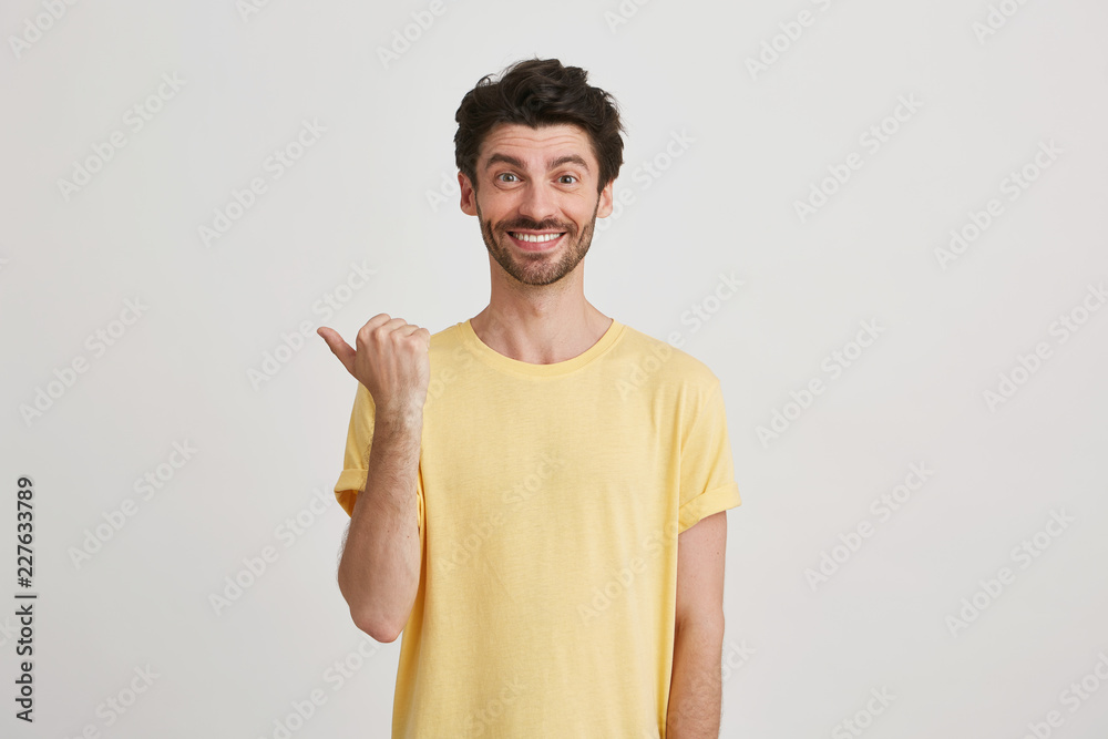 Fototapeta premium Smiling happy attractive young man with dark brown hair and beard wears yellow tshirt, points left with one thumb, isolated over white background