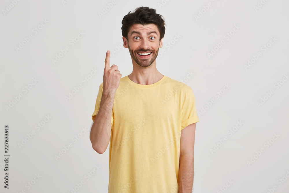 Smiling happy attractive young man with dark brown hair and beard wears yellow tshirt, points up with one finger, looks like having a great idea isolated over white background Smiling happy attractive young man with dark brown hair and beard wears yellow tshirt, points up with one finger, looks like having a great idea isolated over white background