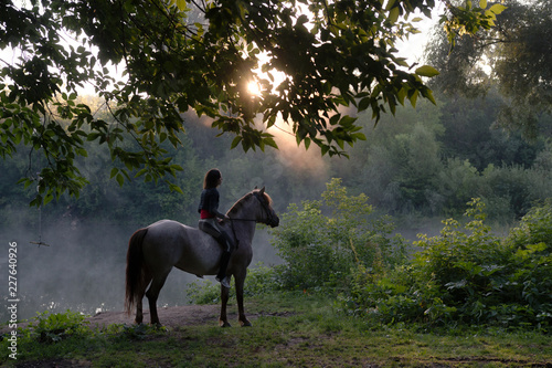 Young woman riding a white horse on a beautiful landscape. Clear lake at morning fog. Sunrise