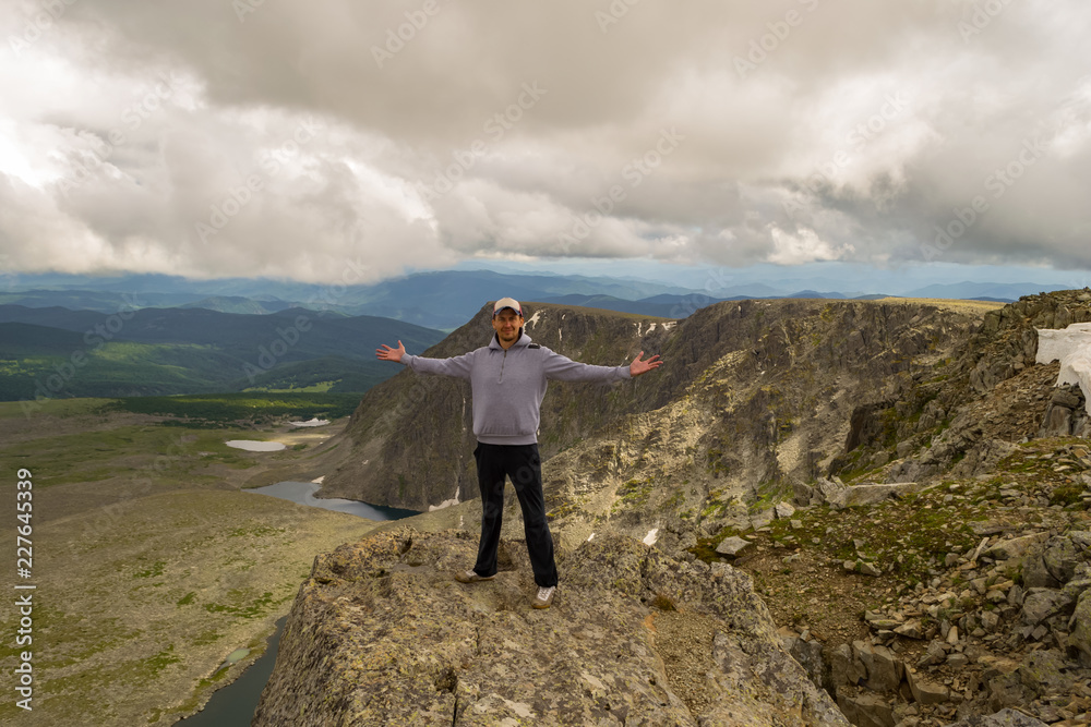 Obraz premium Guy in a gray sweatshirt with a cap is standing with his hands apart on the edge of the cliff in front of him is a colorful view of mountains lakes with glaciers. Traveler complete the route
