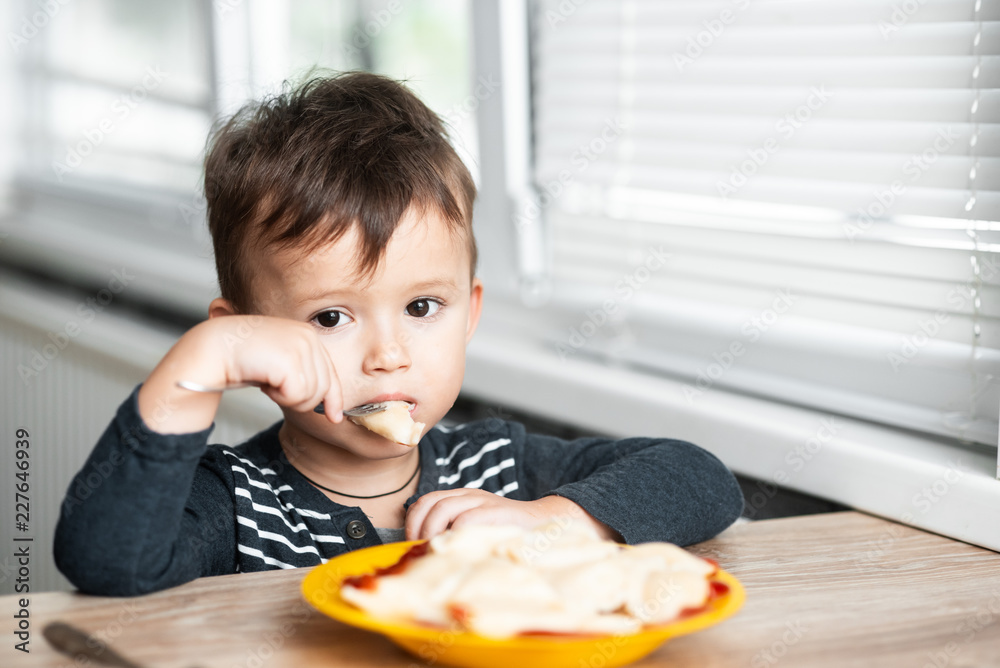 Hungry child eating dumplings in the kitchen, sitting at the table in a gray jacket Hungry child eating dumplings in the kitchen, sitting at the table in a gray jacket