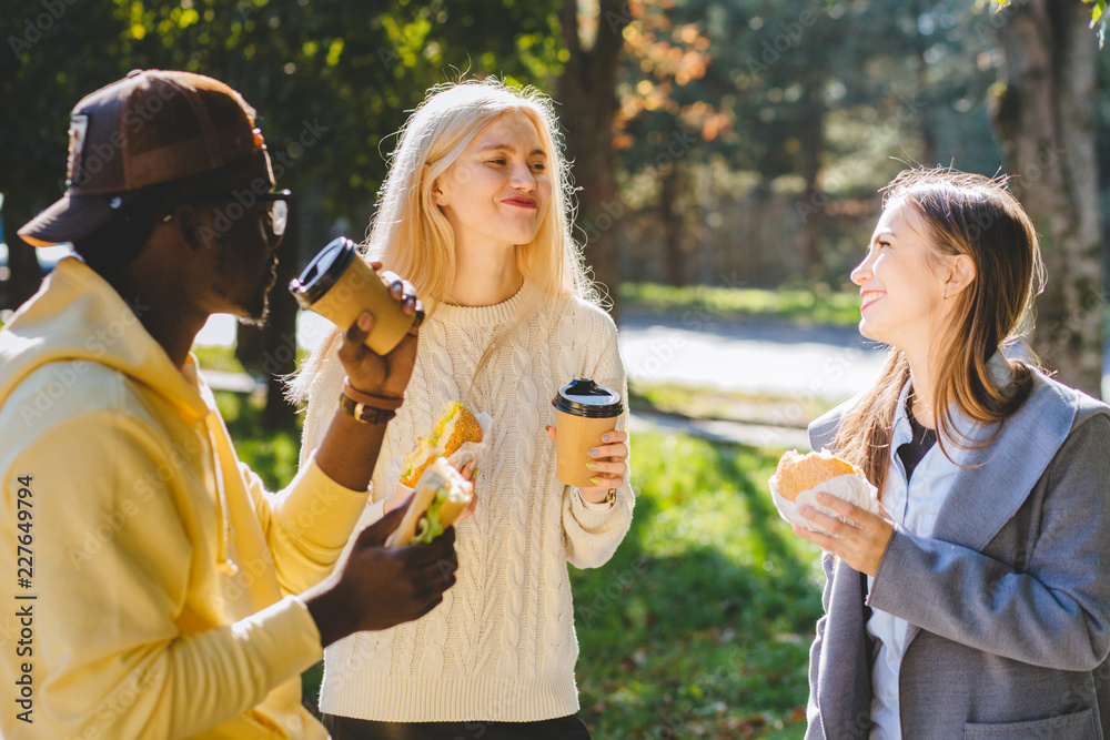 Three young multiethnic hungry people students discussing, having lunch, and eating to-go food while standing up outdoor in sunny autumn day. Making friends, get-together meeting concept.