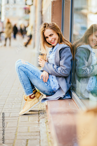 Brunette trendy woman portrait of young pretty trendy girl sitting near shop window, drinking coffee at the city in Europe, autumn street, laughing and smiling portrait.