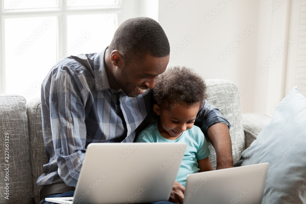 American family spend free time together at home. African father and little toddler son with laptop sits on couch together, teach learn how use pc, watch videos play online games like father like son American family spend free time together at home. African father and little toddler son with laptop sits on couch together, teach learn how use pc, watch videos play online games like father like son