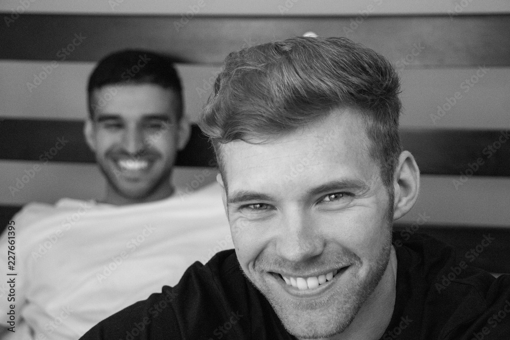 Gay lovers sitting in bed with one close to camera and the other looking on. Both are smiling. Gay lovers sitting in bed with one close to camera and the other looking on. Both are smiling.