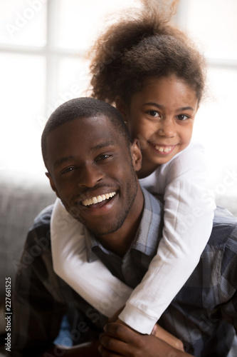 Portrait black African dad and kid smiling laughing looking at camera sitting on couch at home. American attractive daddy with little preschool beautiful daughter. Father day and Happy family concept