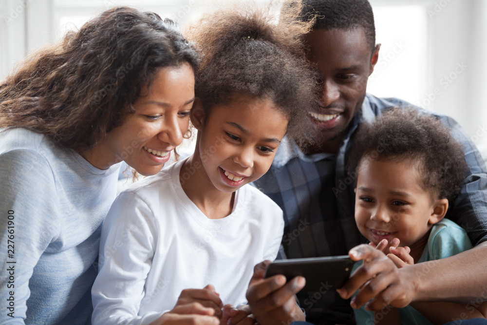 Close up black family with small children have a fun sitting on couch  together at home. Happy american married couple little preschool adorable  daughter and son watching funny video using smartphone Stock
