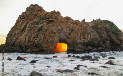 Orange Sunset streams through keyhole arch rock formation in Big Sur