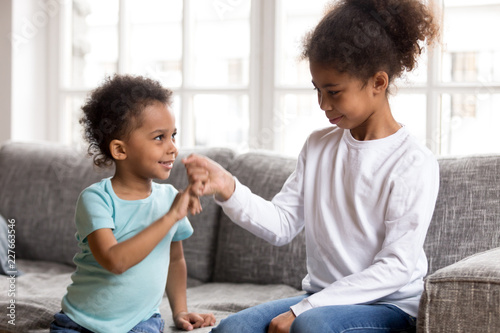 Black African little brother and sister sitting on couch at home. Small american children reconcile after fight or quarrelling making peace with hand gesture joining pinkies swear be friends forever photo