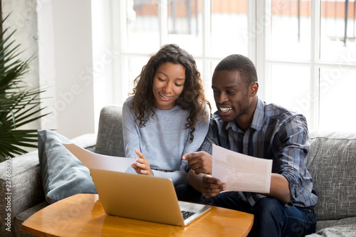 African black positive married couple sitting on sofa at home read documents paper checking bills, bank account balance feeling satisfied and happy. Refund income last loan payment good news concept