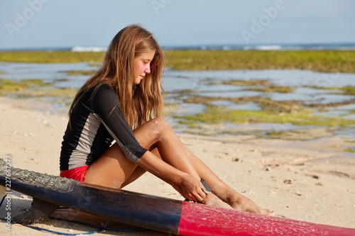 Sideways shot of attractive slim young female surfer fastens leash on leg which allows be save from crashing into rocky shorelines , studies waves before entering water, near surfboard on ocean shore photo