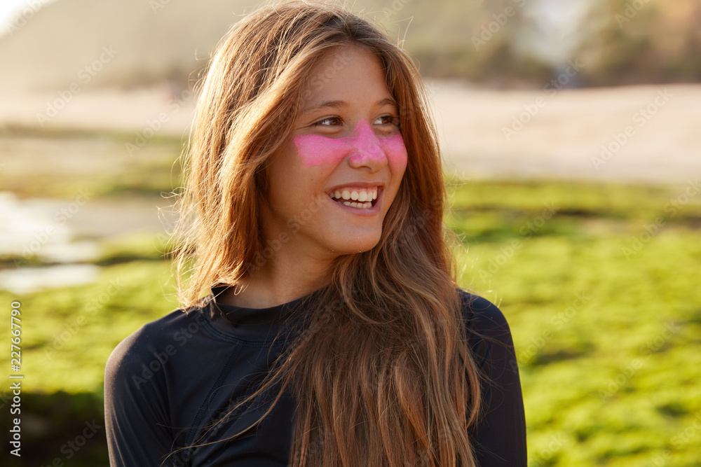 Close up shot of cheerful Caucasian female likes surfing, looks happily aside, has surf zinc on cheeks, poses against blurred outdoor background on seashore. Positive emotions and hobby concept Close up shot of cheerful Caucasian female likes surfing, looks happily aside, has surf zinc on cheeks, poses against blurred outdoor background on seashore. Positive emotions and hobby concept