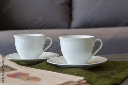 Two coffee cups put on plate and dark green cloth with newspaper in the living room and window lighting. Selective focus.