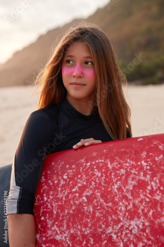 Perfect sunny day for surfing. Contemplative surfer in wetsuit, has healthy skin, protects face with surf zinc, waits for big waves. Female amateur of sport and extreme activities, poses on coastline photo