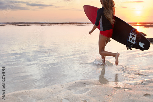 Back view of running girl surfer in swimsuit, carries board under arm, ready to conquer giant wave, runs into ocean, beautiful panoramic view, sunset, wet sand with left footrpint on surface
