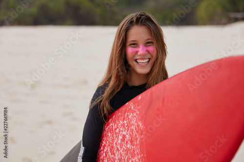 Water sport concept. Happy delighted surfboarder in diving suit, uses red board for hiting waves, being in good mood, poses against blurred background outdoor. Tourist during holiday in exotic country photo