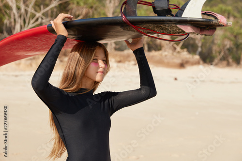 Photo of self assured professional woman wears beaver tail, waits for big surfs, stands at bone yard, being goofy foot, ready to hit surf, poses against sandy beach, afraids of rip, enjoys spare time