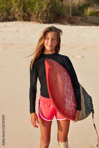 Fit active healthy surfer gazes into distance with pensive expression, stands with surfboard on sandy exotic beach, involved in watersports, wears divingsuit. Recreational activity and hobby concept photo