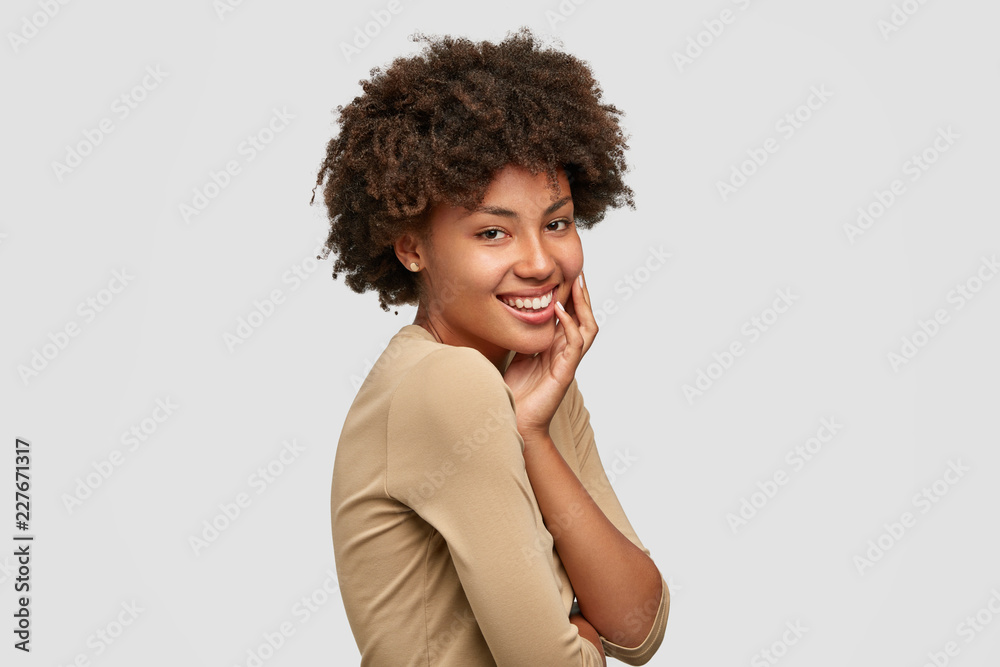Sideways shot of cheerful dark skinned young model with Afro haircut, has gentle smile, dressed in casual beige sweater, models in studio against white background. People and ethnicity concept Sideways shot of cheerful dark skinned young model with Afro haircut, has gentle smile, dressed in casual beige sweater, models in studio against white background. People and ethnicity concept