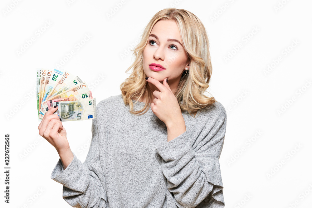Young thinking pretty woman in grey sweater holding bunch of Euro banknotes, looking upwards with hand on chin, isolated on white background. Young thinking pretty woman in grey sweater holding bunch of Euro banknotes, looking upwards with hand on chin, isolated on white background.