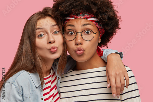 Close up shot of appealing beautiful mixed race females make grimace at camera, pout lips as going to kiss someone, cuddle with each other, demonstrates good relationships, isolated on pink wall