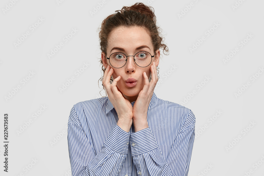 Surprised Caucasian woman with freckled skin, wears optical glasses, keeps hands on cheeks, has unbelievable look, dressed in striped shirt, isolated over white background. Facial expressions concept