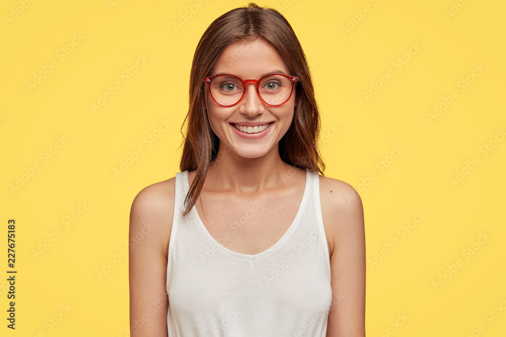 Horizontal view of cheerful emotive brunette girlfriend in optical glasses and white vest, has positive smile on face models over yellow background. Satisfied journalist has great news for publication Horizontal view of cheerful emotive brunette girlfriend in optical glasses and white vest, has positive smile on face models over yellow background. Satisfied journalist has great news for publication