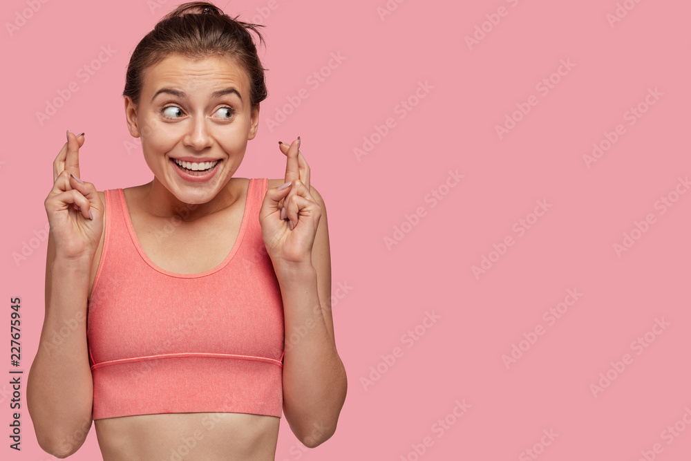 Horizontal shot of pleased young woman with sporty body, crosses fingers for making wish, has satisfied facial expression, models against pink background with free space for your advertisement Horizontal shot of pleased young woman with sporty body, crosses fingers for making wish, has satisfied facial expression, models against pink background with free space for your advertisement