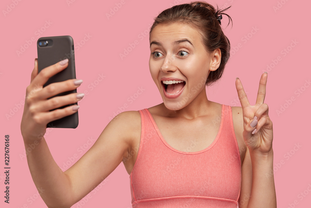 Horizontal view of cheerful European woman makes peace gesture at camera of cell phone, dressed in pink vest, being in good mood, shares photos in social networks, poses for selfie indoor, isolated Horizontal view of cheerful European woman makes peace gesture at camera of cell phone, dressed in pink vest, being in good mood, shares photos in social networks, poses for selfie indoor, isolated